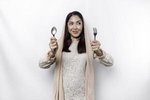 An Asian Muslim woman is fasting and hungry and holding utensils cutlery while looking aside thinking about what to eat photo