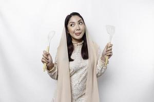 Excited Asian Muslim woman wearing a headscarf, holding cooking ware and smiling, isolated by white background photo