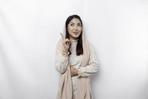 Starving Asian Muslim woman wearing a headscarf, pointing up at the copy space above her, isolated by a white background photo