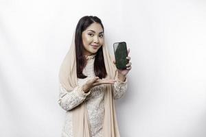 A portrait of a happy Asian Muslim woman wearing a headscarf, showing her phone screen, isolated by white background photo