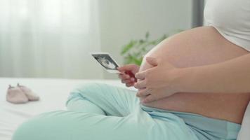 zwanger vrouw is op zoek Bij een echografie foto van foetus. moeder voorzichtig accenten de baby Aan maag. Vrolijk, familie, groei, zwangerschap, genot , bereiden pasgeboren, nemen zorg, gezondheidszorg, buik. video
