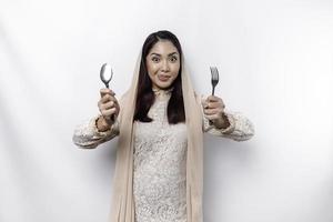 An Asian Muslim woman is fasting and hungry and holding utensils cutlery while looking aside thinking about what to eat photo