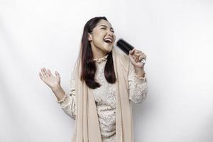 Portrait of carefree Asian Muslim woman, having fun karaoke, singing in microphone while standing over white background photo