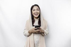 A portrait of a happy Asian Muslim woman wearing a headscarf, holding her phone, isolated by white background photo