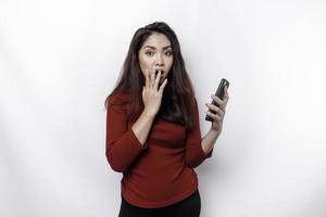 Shocked Asian woman wearing red top, holding her phone, isolated by white background photo