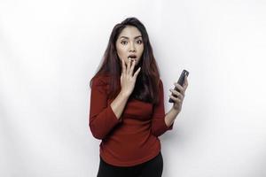 Shocked Asian woman wearing red top, holding her phone, isolated by white background photo