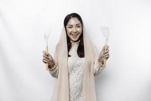 Excited Asian Muslim woman wearing a headscarf, holding cooking ware and smiling, isolated by white background photo