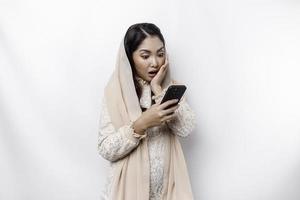 Shocked Asian woman wearing headscarf, holding her phone, isolated by white background photo