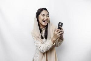 A portrait of a happy Asian Muslim woman wearing a headscarf, holding her phone, isolated by white background photo