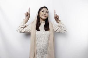 Excited Asian Muslim woman wearing hijab pointing at the copy space above her, isolated by white background photo