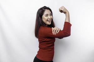 emocionado asiático mujer vistiendo un rojo parte superior demostración fuerte gesto por levantamiento su brazos y músculos sonriente con orgullo foto