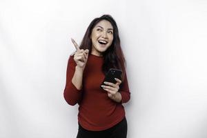 Excited Asian woman wearing red top pointing at the copy space on top of her while holding her phone, isolated by white background photo