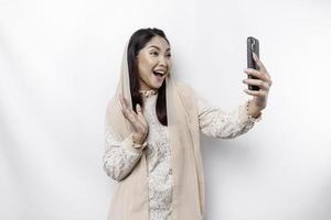 un retrato de un contento asiático musulmán mujer vistiendo un Pañuelo, participación su teléfono, aislado por blanco antecedentes foto