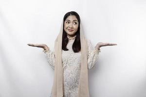 A thoughtful young Asian Muslim woman is wearing hijab and looks confused between choices, isolated by a white background photo