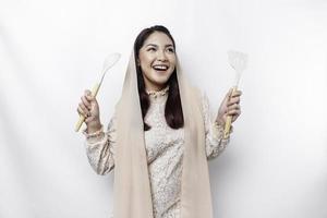 Excited Asian Muslim woman wearing a headscarf, holding cooking ware and smiling, isolated by white background photo