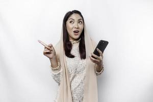 Shocked Asian Muslim woman wearing hijab pointing at the copy space beside her while holding her phone, isolated by white background photo