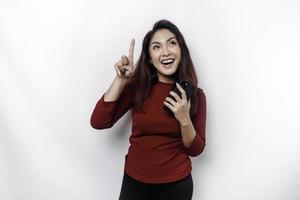 Excited Asian woman wearing red top pointing at the copy space on top of her while holding her phone, isolated by white background photo