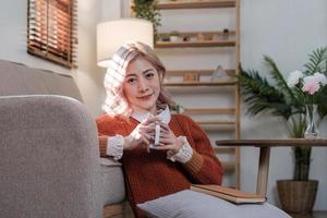 Young beautiful Asian woman relaxing in living room at home, reading books, drinking coffee and using phone in the morning with happy feeling photo