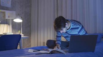 adolescente estudiando en cama a noche y haciendo tarea. joven hombre estudiando a noche en su cama. allí son libros y ordenador portátil en frente de a él. video