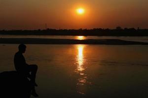 silueta de un hombre sentado en un rock por el río mirando a puesta de sol foto