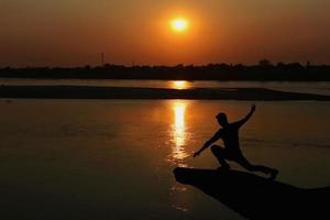 silueta de un hombre hacer yoga en el banco de el río a puesta de sol. sano concepto. foto