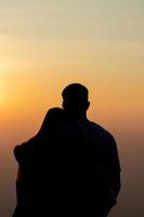 silueta de un joven Pareja acecho el puesta de sol durante crepúsculo en un alto montaña y abrazando felizmente. contento Pareja abrazando cada otro con amor y amistad y amabilidad para cada otro. foto