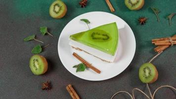 Close up of Homemade Cheesecake with Kiwi in a white plate on a Green background. Decoreted with slice of kiwi, cinnamon stick and leaves mint