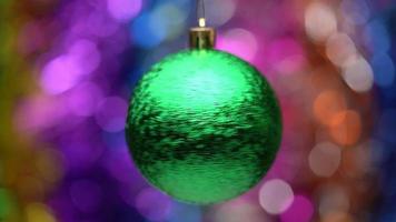 Spinning New Year's ball green color on blurry colored bokeh background of glowing tinsel, holiday lights. Close up of hanging holiday Christmas ball. Soft, selective focus on foreground. Motion blur video