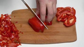 Close up view of Caucasian male cook chopping red bell pepper with sharp knife into small slices on wooden kitchen cutting board. Slicing sweet pepper, work process of cooking vegetables in kitchen video