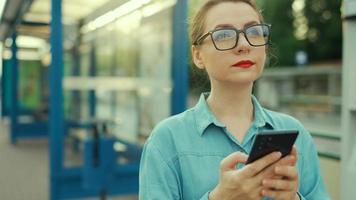 mujer soportes a un transporte detener, utilizando teléfono inteligente y esperando para el tranvía. video