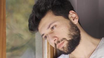 depresso uomo guardare su il finestra pensieroso. giovane uomo con psicologico i problemi è infelice e sembra impotente su di il finestra di il Casa. video