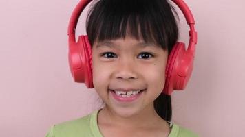 linda pequeño niña en auriculares escucha a música. inalámbrico auriculares dispositivo accesorio. niño disfrutando música en auriculares en pastel rosado antecedentes en estudio. video