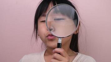 Little girl looking through a magnifying glass on pastel pink background in studio. Curious schoolgirl in casual clothes looking at the camera through a magnifying glass. video