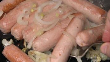 Closeup view of hands chef stirring sausages with metal fork. Stirs and cooking appetizing sausages with onion fried in black pan video