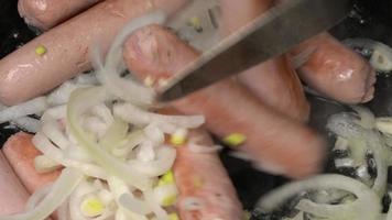 Cooking and stirring sausages with onion fried in black pan. Hands chef stirs sausages with metal spatula. Close-up view video