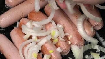 Cook adds onions to sausages and is fried in black pan with oil. Close-up view of cooking of delicious, high-calorie junk food with large content of cholesterol. video