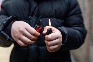 Boy lighting up several firecrackers in his hand using lighter. Kid getting ready for celebration with fireworks or pyrotechnic products . Boy holding a burning petard in his hand. Close up shot photo