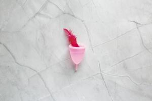 Menstrual cup and red bird feather on marble background. Alternative feminine hygiene product during the period. Women health concept photo