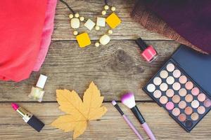 Women's clothing and cosmetics sweaters, lipstick, nail polish, necklaces, eye shadow, brushes, yellow leaves on wooden background. Top view. Toned image. photo