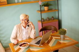 Senior woman using mobile phone while working on laptop and having healthy breakfast in the cafe photo