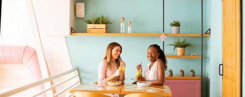 Young black and caucasian woman having good time, drinking fresh juices and having healthy breakfast in the cafe photo