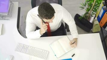 Businessman talking on phone in office and taking notes in notebook. Businessman talks on the phone at his desk and gets things done by taking notes. video