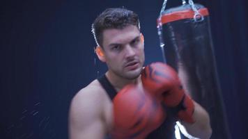 agresivo masculino Boxer profesionalmente boxeo en un oscuro gimnasia. masculino kickboxer lanza su puños hacia el cámara y esquiva puñetazos con su guantes. sombra boxeo. video