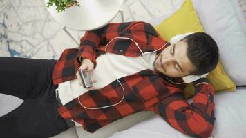 Young man listening to music with headphones resting on comfortable sofa and happy. Relaxing young man sitting on sofa at home listening to music and being happy. video