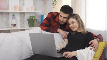 feliz jovem casal compras conectados juntos usando computador portátil para comércio eletrônico. moderno jovem casal sentado em sofá às casa em repouso, relaxante olhando às computador portátil compras on-line, assistindo filmes. video