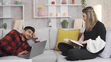 Happy young couple sitting on sofa at home, having a pleasant and intimate time. Young man looking at laptop and talking to wife reading book, they are happy and cute. video