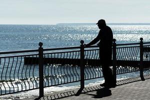 Aged man dreams about life adventures, past traveling and looks at sea. Rear back view, copy space. Old cancer widower misses his wife. Sunny weather and clean blue sea. photo