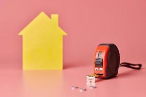 House renovation funny concept. Metal tape measure and other repair items. Home repair and redecorated concept. Yellow house shaped figure on pink background. photo