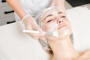 Beautician applies vitamins cream mask on woman face for rehydrate face skin in beauty salon photo
