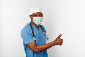 negro cirujano médico hombre en azul Saco blanco gorra y cirujano máscara con pulgares arriba gesto foto